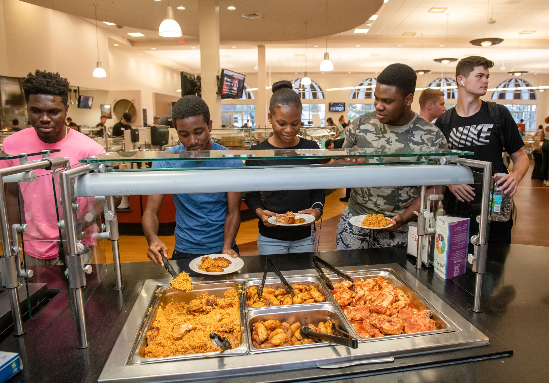 Four students, two men and two women, selecting food at a buffet line in a college dining hall